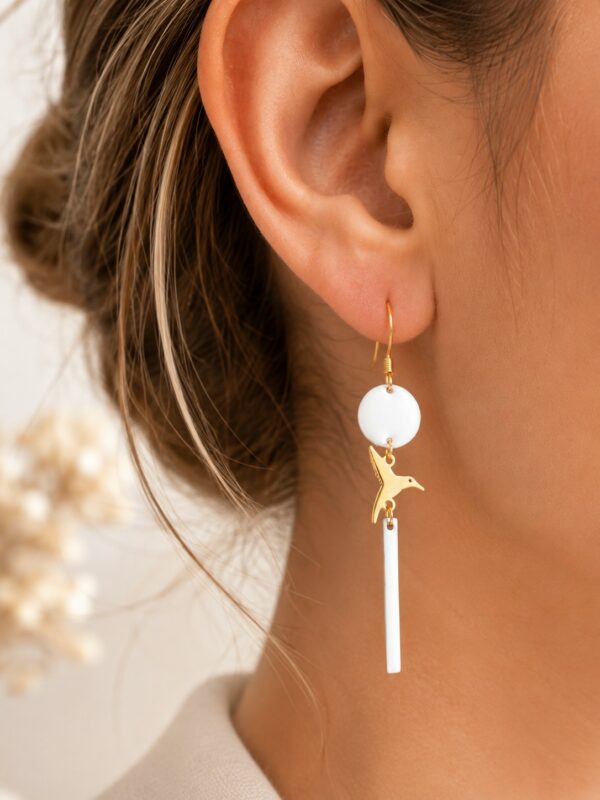 Boucles d'oreilles Alba pendantes dorées et blanches avec motif oiseau portées sur une oreille.