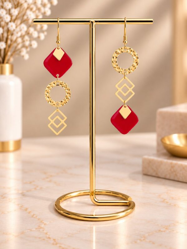 Boucles d'oreilles Pandore asymétriques dorées et rouges présentées sur support.