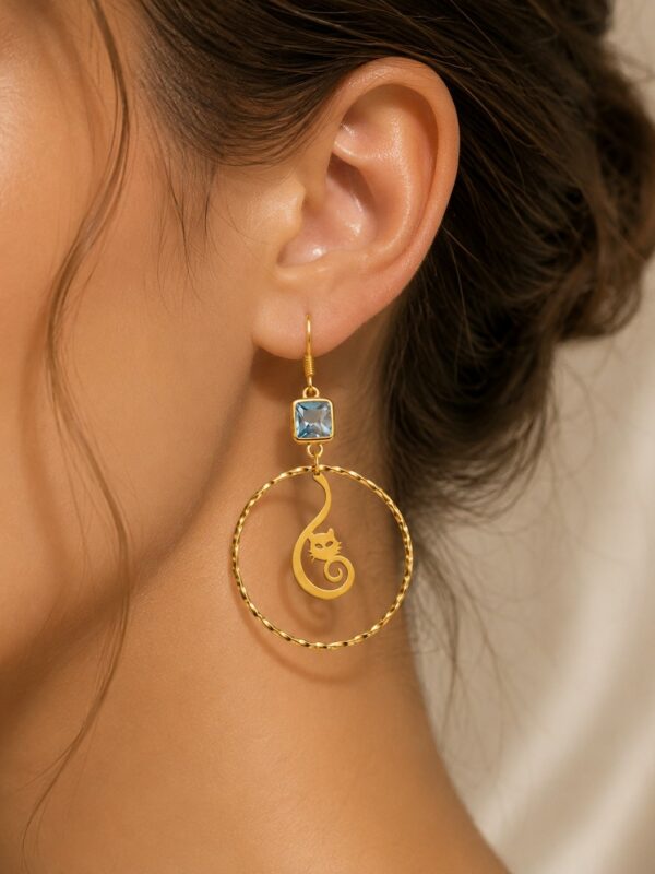 Boucles d'oreilles Clara pendantes dorées et bleues motif chat portées sur une oreille.